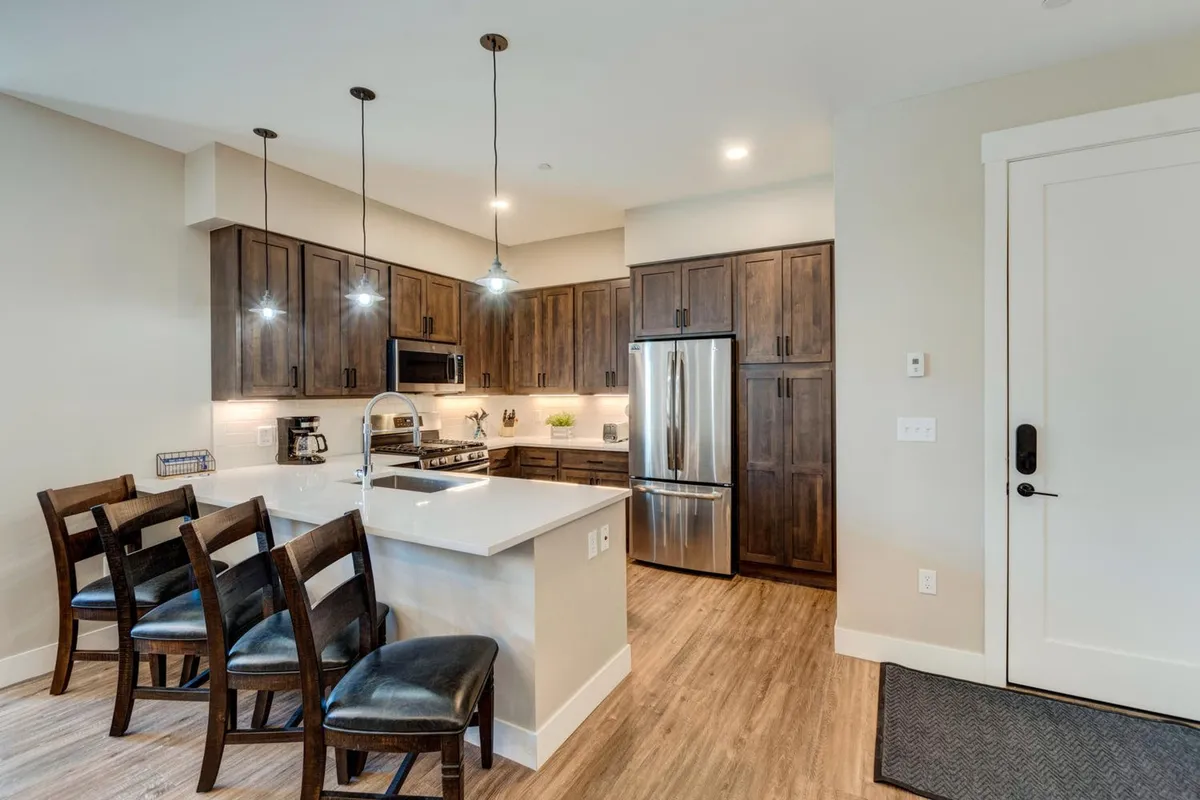 A modern kitchen with a prominent island, sleek appliances, wood cabinetry, and bar stools. The scene suggests a space designed for communal cooking, entertaining, and casual dining.