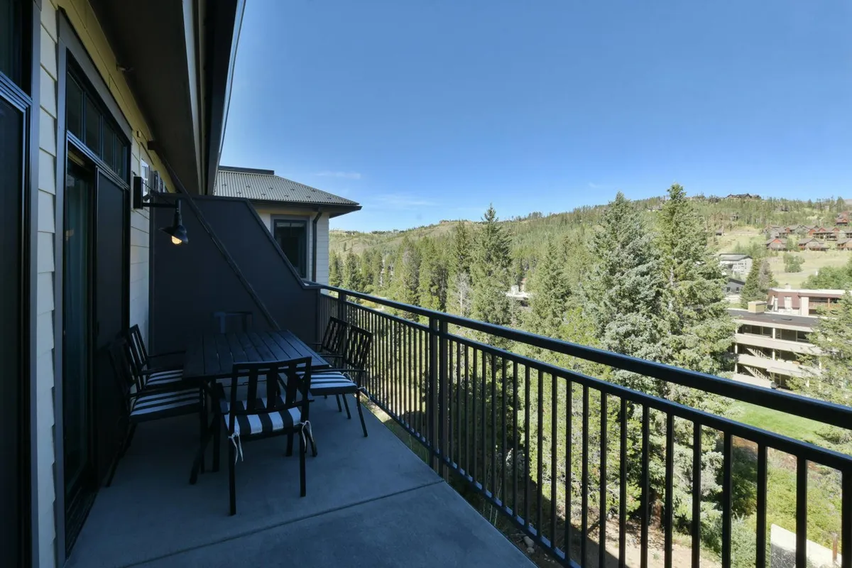 This image features a balcony with a railing, a table and chairs for seating, surrounded by trees and with sweeping mountain views in the background. The setup invites relaxation or casual dining, and the natural environment is tranquil and picturesque.