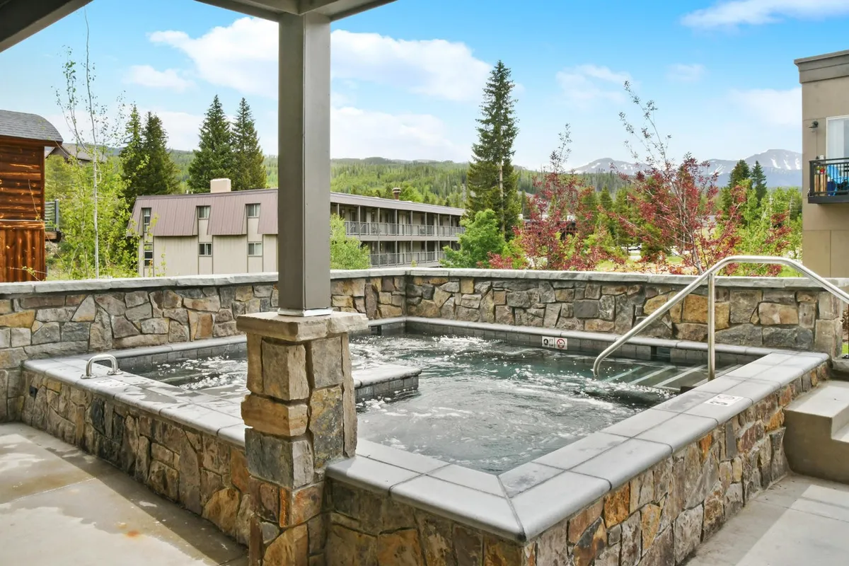 An outdoor hot tub set against a stone wall, surrounded by lush greenery and offering expansive views of the mountains. The scene suggests a tranquil space perfect for socializing and relaxing after a day of group activities, with enough room for multiple people to enjoy together.