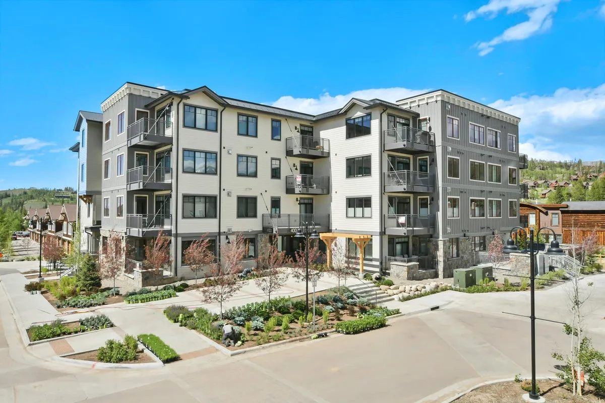 A wide view of a modern building with multiple balconies and landscaped gardens, where people can be seen relaxing or gathering outdoors. The architecture and outdoor spaces blend contemporary comfort with an inviting, social vibe, and the balconies offer mountain views.