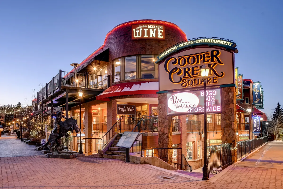 This exterior scene captures Cooper Creek Square with its festive lighting, visible shop and wine bar signs, and outdoor seating area. Decorative lighting and signage suggest bustling activity and a lively, inviting atmosphere for shopping and dining downtown.
