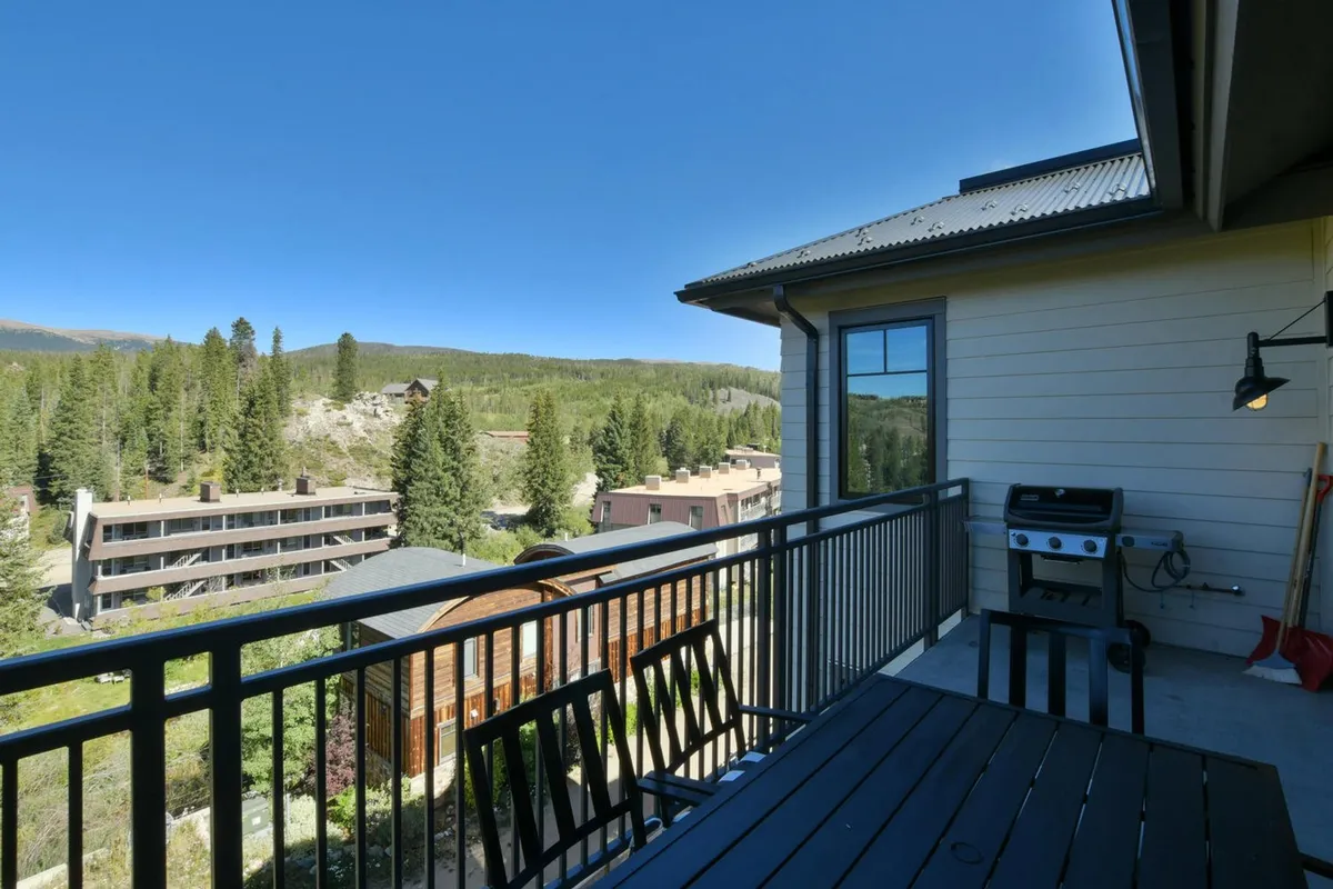 An exterior view of a balcony with a grill, a wooden dining table, comfortable seating, and abundant greenery with a stunning mountain backdrop. The image highlights ample space for outdoor dining and gatherings, along with the presence of a grill for barbecuing.
