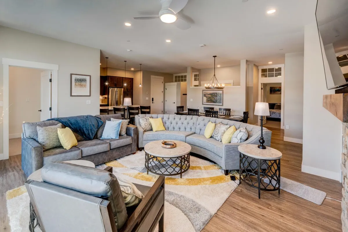 An open, inviting living area featuring a large sectional sofa, a coffee table, a rug, and a view into the adjacent dining area. The space is softly lit, evoking warmth and comfort, perfect for gatherings, movies, or game nights.