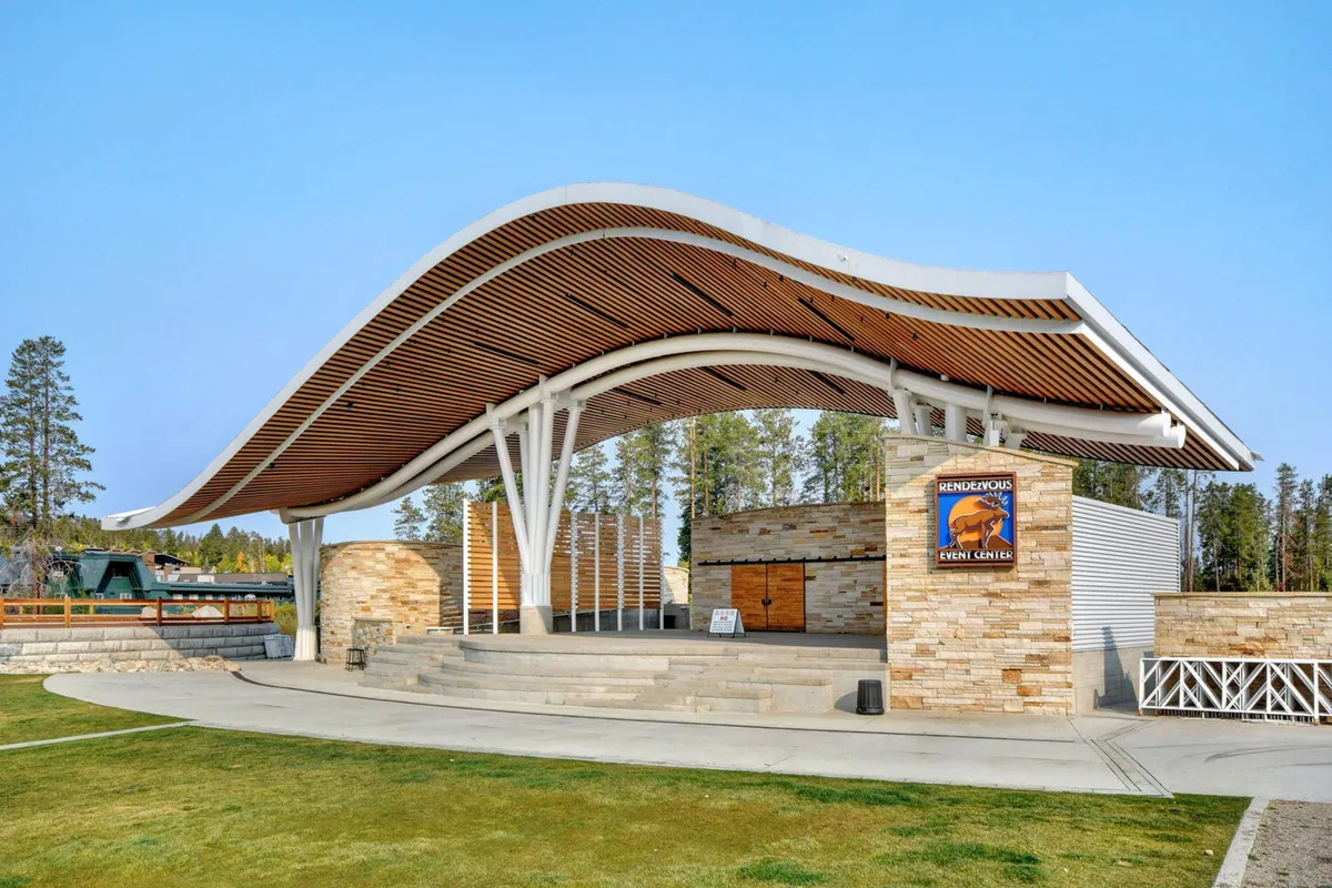 An exterior shot of a distinctive event center with a curved roof and stone façade, featuring prominent signage and a lively, welcoming atmosphere. Visible elements suggest preparations for or hosting of concerts, festivals, or community gatherings, with open spaces ideal for outdoor events.