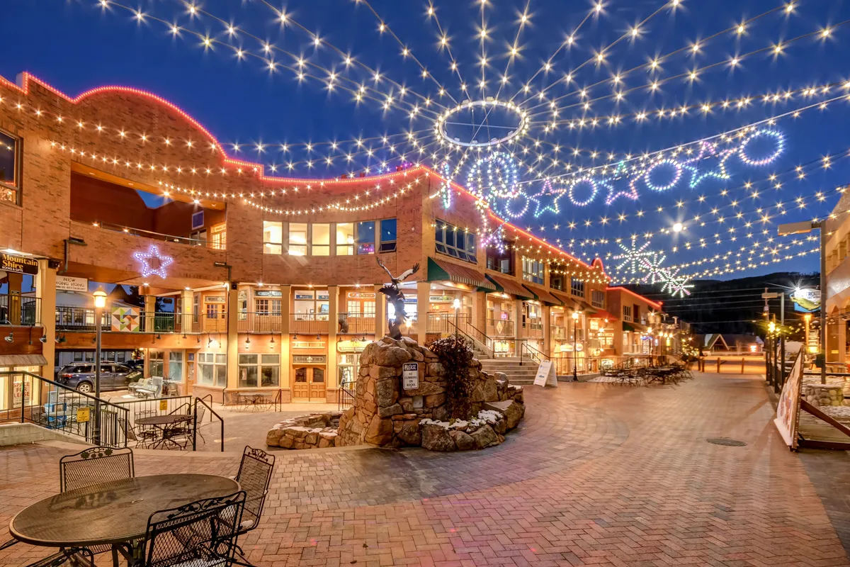 A festive town square scene illuminated by string lights and decorative stars, with a stone fountain at the center and surrounded by local boutiques and eateries. People are seen dining al fresco, shopping, and enjoying the vibrant community atmosphere, capturing both day and evening activity.