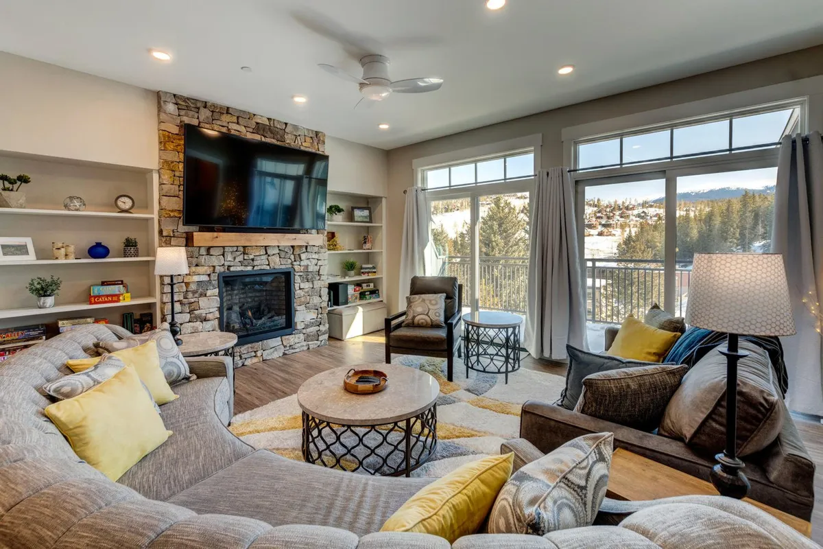 The image showcases a spacious, inviting living room with a stunning stone fireplace as the centerpiece. Floor-to-ceiling windows reveal sweeping mountain views, while a plush curved sectional sofa and coffee table create a perfect spot for gathering and relaxing. The scene exudes warmth and comfort, ideal for cozy nights in after adventure-filled days in Winter Park.