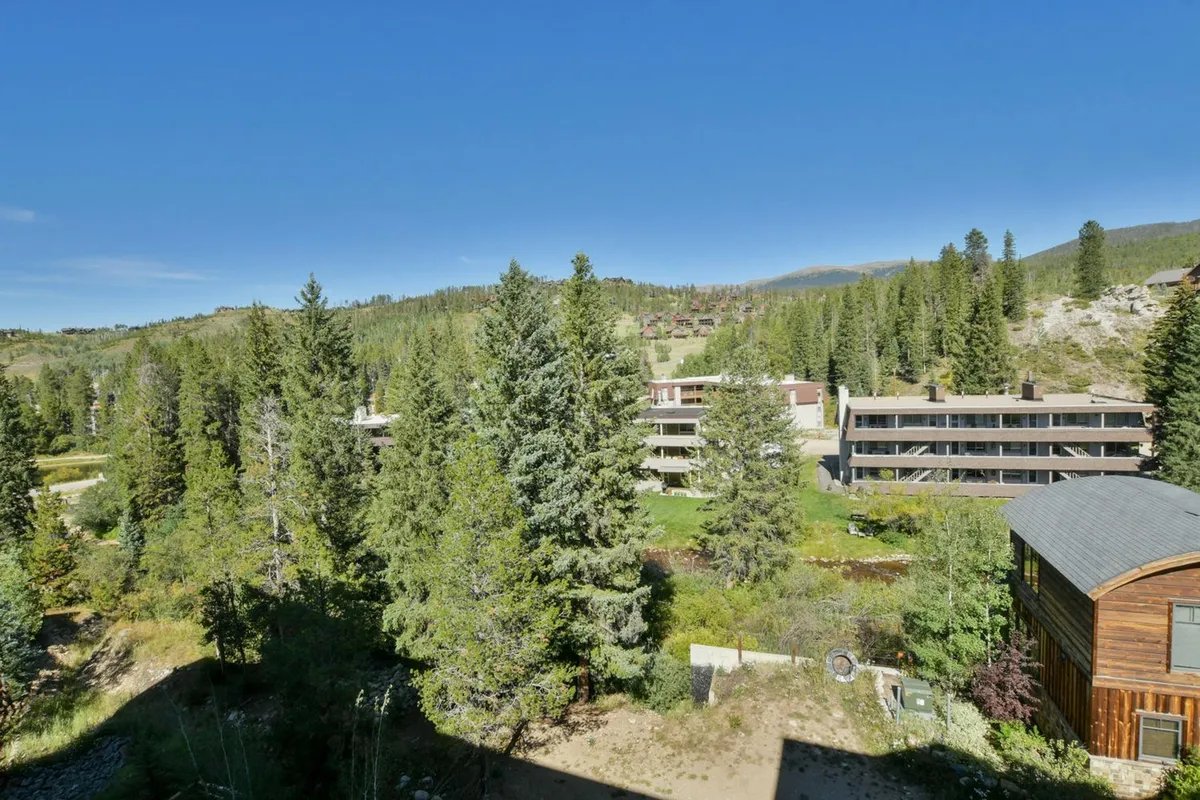 This image showcases tall pine trees framing modern condo buildings with distant mountains visible in the background. The scene is tranquil and highlights the property's natural setting, suggesting proximity to trails and outdoor recreation areas ideal for dog walks and adventure.